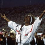 Whitney Houston sings with her arms outstretched in a tracksuit and white headband at the superbowl