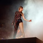 Taylor Swift on stage with a black unitard and red applique with fog behind her