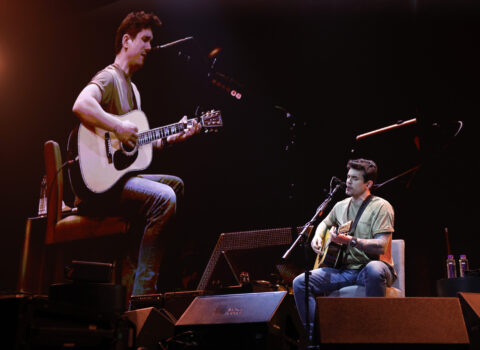 John Mayer performing on stage while seated with a projection of himself on the screen behind him