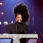 Janet Jackson behind a podium with two trophies in front of her wearing all black with her hair poofed up
