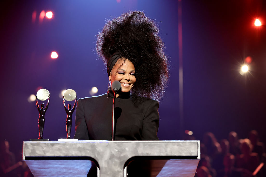 Janet Jackson behind a podium with two trophies in front of her wearing all black with her hair poo...