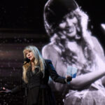 Stevie Nicks wears all black performing in front of a microphone with an image of her younger self behind her