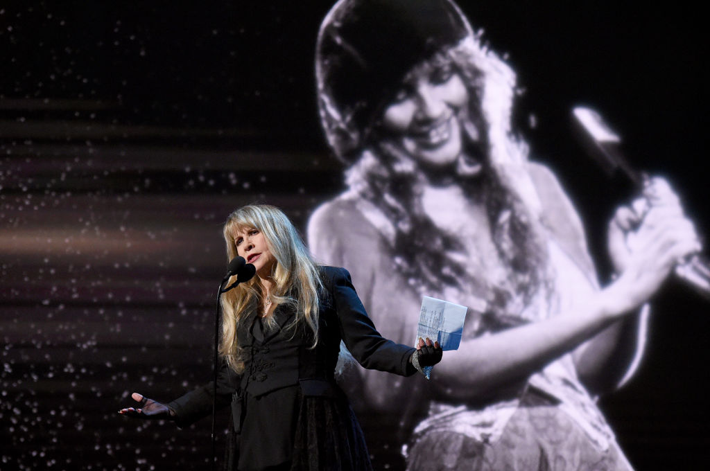 Stevie Nicks wears all black performing in front of a microphone with an image of her younger self behind her
